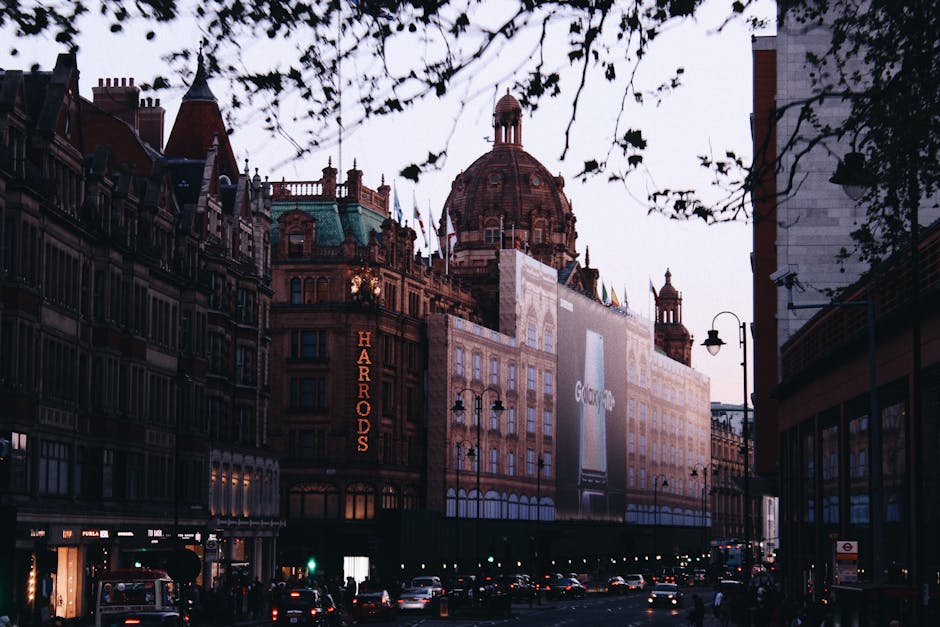 A city street scene in front of the Harrods storefront in Knightsbridge during early evening, with the iconic domed building visible in the background. The street is lined with historic, ornate buildi