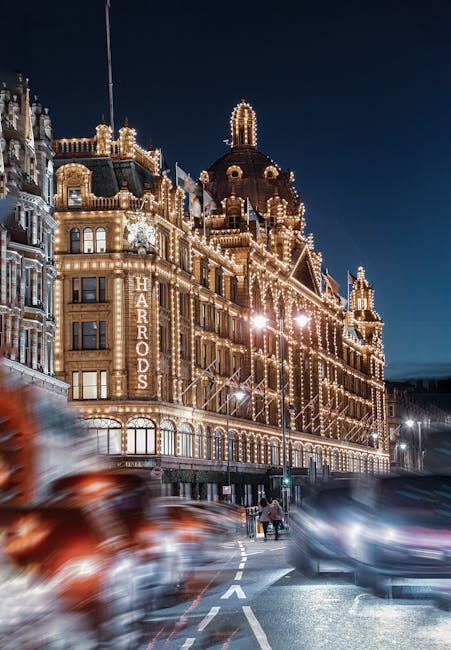 A city street scene in front of the Harrods storefront in Knightsbridge during early evening, with the iconic domed building visible in the background. The street is lined with historic, ornate buildi