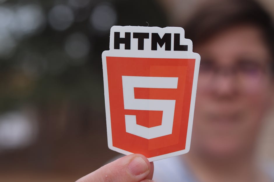 A person holding a sticker or sign displaying the HTML5 logo, which features a shield-shaped design with a prominent white number '5' on an orange background, and the text 'HTML' in black at the top.