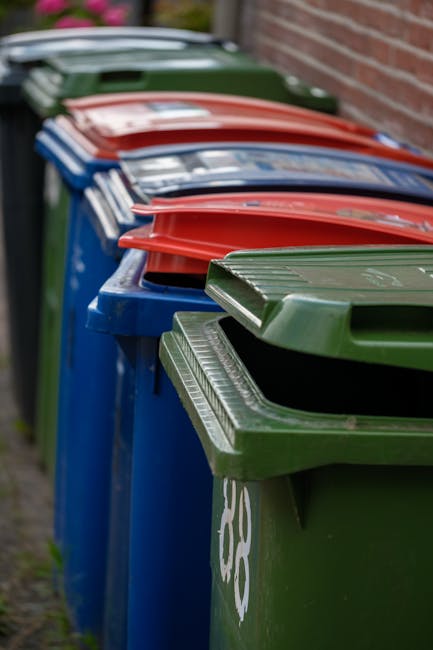 A row of outdoor garden waste bins lined up along a narrow paved pathway adjacent to a brick wall. The bins are color-coded with green, blue, red, and black lids, each labeled for different types of g