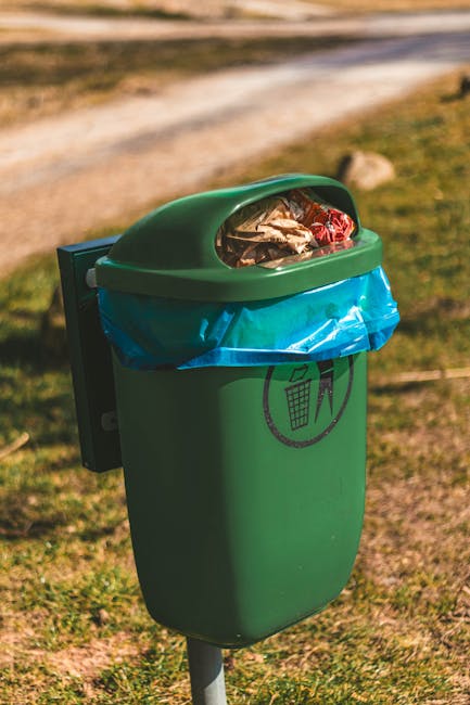 A row of outdoor garden waste bins lined up along a narrow paved pathway adjacent to a brick wall. The bins are color-coded with green, blue, red, and black lids, each labeled for different types of g